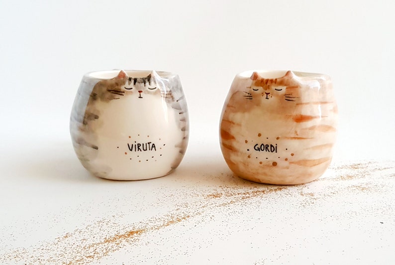 May include: Two ceramic cat mugs with painted details. The gray mug has the name "Viruta" written on it and the brown mug has the name "Gordi" written on it.