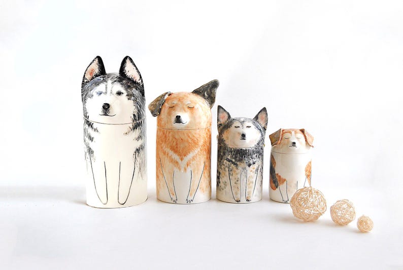 May include: Four ceramic dog figurines, each with a different breed and color. The figurines are arranged in a row from largest to smallest. The largest figurine is a black and white husky, followed by a brown and white dog, then a black and white dog, and finally a brown and white dog.