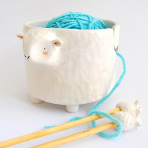 May include: White ceramic yarn bowl shaped like a sheep with a blue ball of yarn inside. The bowl has a small hole for the yarn to feed through. Two wooden knitting needles are resting on the yarn.