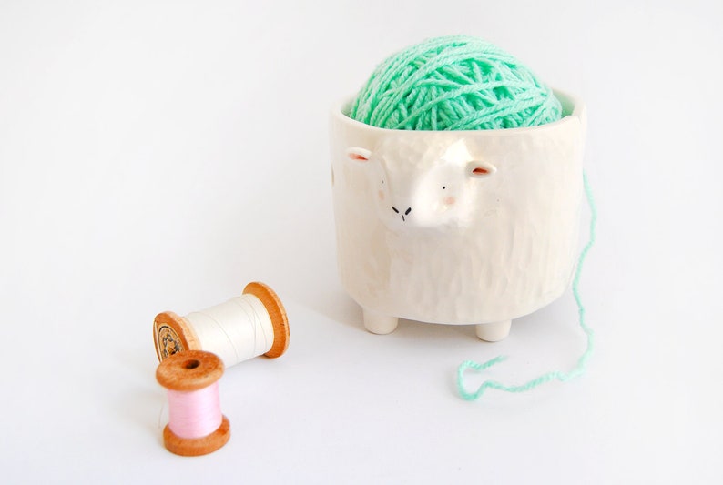 May include: A white ceramic sheep yarn bowl with a ball of light green yarn inside. Two spools of thread, one white and one pink, are on the white surface.