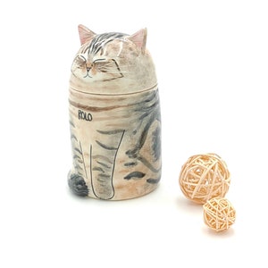 May include: A ceramic cat figurine with a closed-eye expression and the name "ROLO" written on its side. The figurine is sitting on a white surface with two small, woven balls beside it.