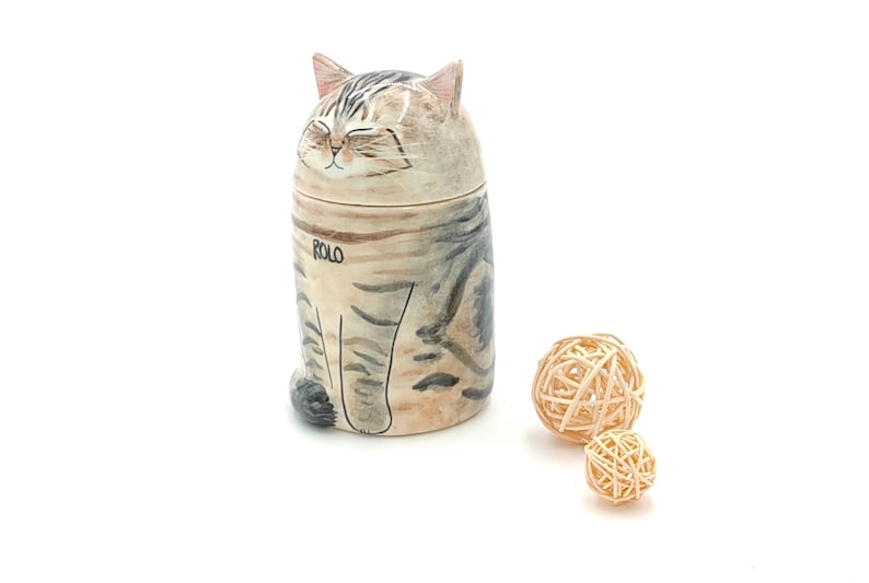 May include: A ceramic cat figurine with a closed-eye expression and the name "ROLO" written on its side. The figurine is sitting on a white surface with two small, woven balls beside it.