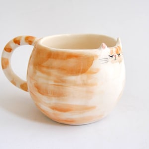 May include: A ceramic mug shaped like a cat with a white body and orange stripes. The mug has a handle and a sleepy cat face with closed eyes.