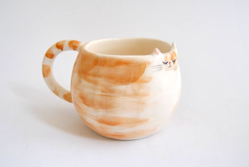 May include: A ceramic mug shaped like a cat with a white body and orange stripes. The mug has a handle and a sleepy cat face with closed eyes.