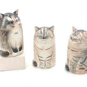 May include: Three ceramic cat figurines, each with a different expression. The cat on the left is a calico cat with the name "Kali" written on its belly. The middle cat is a tabby cat with blue eyes. The cat on the right is a tabby cat with its eyes closed.