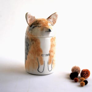 May include: A ceramic pet jar shaped like a fox with closed eyes. The jar is painted in shades of brown, white, and gray. The words "Pet Name" are written on the front of the jar.