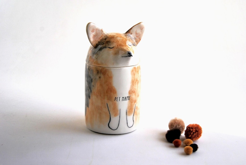 May include: A ceramic pet jar shaped like a fox with closed eyes. The jar is painted in shades of brown, white, and gray. The words "Pet Name" are written on the front of the jar.
