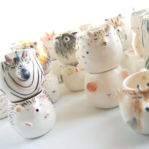 May include: A collection of ceramic animal planters in various colors and designs. The planters are stacked on top of each other and feature animal faces, including a zebra, a hedgehog, a sheep, a fox, and a tiger.