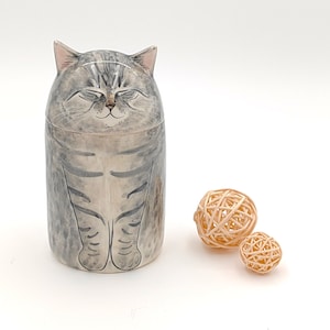 May include: A ceramic cat figurine with a gray and white striped pattern. The cat has closed eyes and a smiling expression. The figurine is sitting on a white surface with two small, woven balls.