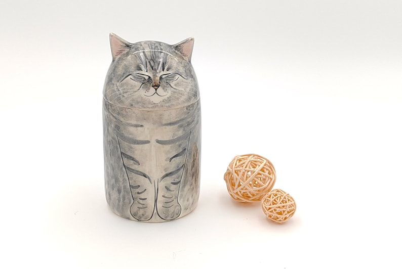 May include: A ceramic cat figurine with a gray and white striped pattern. The cat has closed eyes and a smiling expression. The figurine is sitting on a white surface with two small, woven balls.