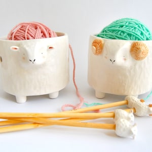 May include: Two ceramic yarn bowls shaped like sheep, one with pink yarn and the other with green yarn. The bowls have small legs and are sitting on a white surface. There are four wooden knitting needles with sheep-shaped ends.