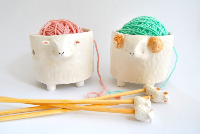May include: Two ceramic yarn bowls shaped like sheep, one with pink yarn and the other with green yarn. The bowls have small legs and are sitting on a white surface. There are four wooden knitting needles with sheep-shaped ends.