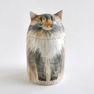 May include: A ceramic cat-shaped jar with a white, gray, and brown painted design. The cat has closed eyes and a fluffy appearance.