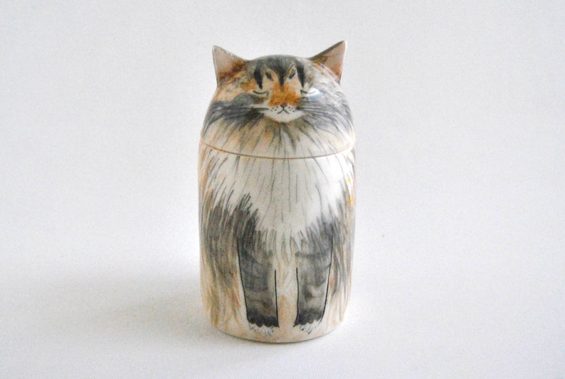 May include: A ceramic cat-shaped jar with a white, gray, and brown painted design. The cat has closed eyes and a fluffy appearance.