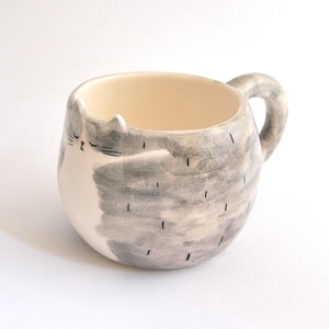 May include: A ceramic mug shaped like a gray and white cat with a closed eye. The mug has a handle and is perfect for holding a warm beverage.
