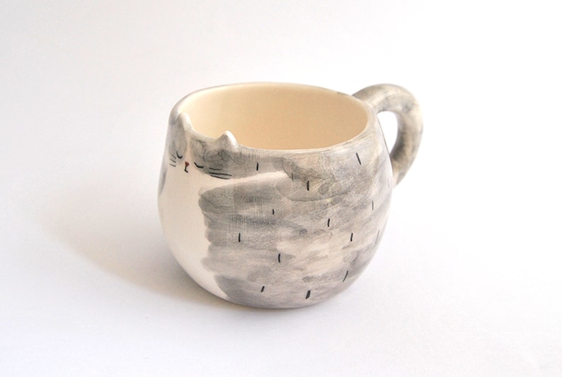 May include: A ceramic mug shaped like a gray and white cat with a closed eye. The mug has a handle and is perfect for holding a warm beverage.
