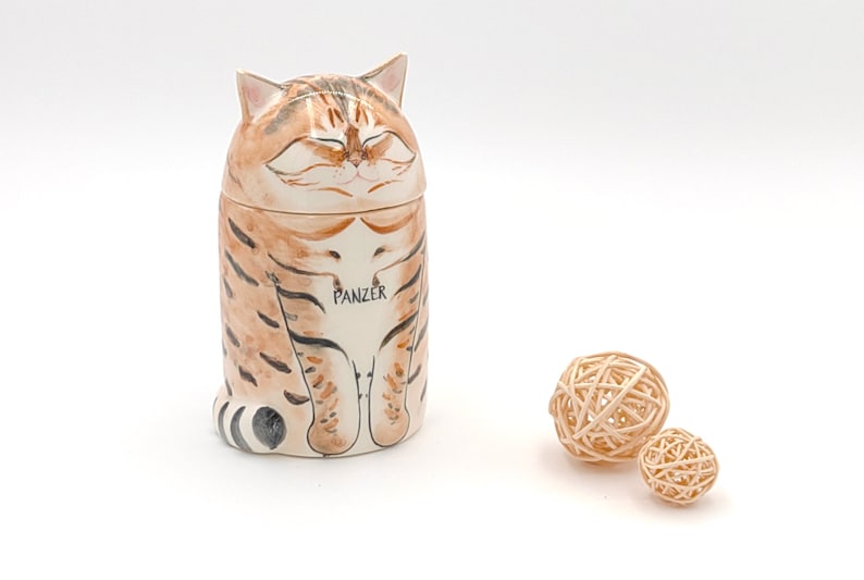 May include: Ceramic cat-shaped container with a brown and white striped pattern. The cat has closed eyes and a relaxed expression. The container is labeled "PANZER".