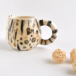 May include: A ceramic mug shaped like a cat with a black and white striped pattern and brown and gold spots. The mug is on a white surface.