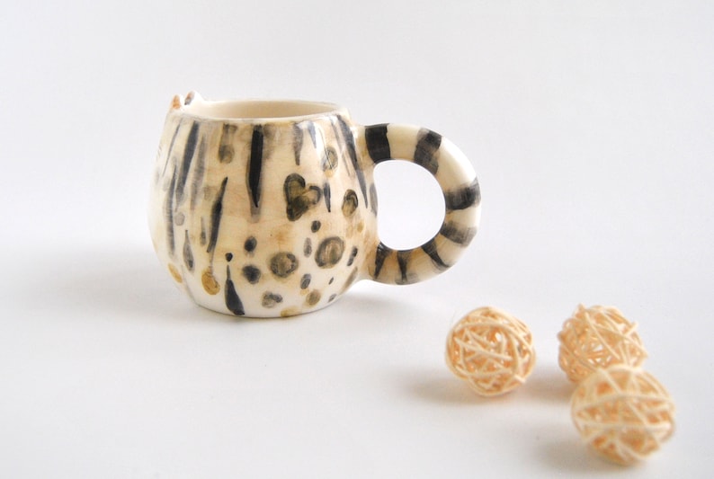 May include: A ceramic mug shaped like a cat with a black and white striped pattern and brown and gold spots. The mug is on a white surface.