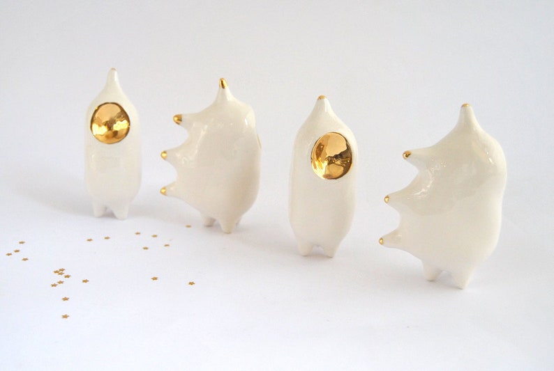 May include: Four white ceramic figurines with gold accents. Each figurine has a different shape and a gold circle on the front. The figurines are arranged in a row on a white surface.
