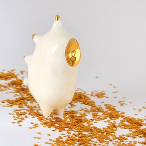 May include: White ceramic sculpture with gold accents, resembling a stylized animal with a rounded body and spiky features. The sculpture is sitting on a white surface covered in gold glitter.