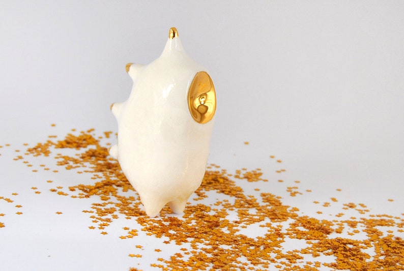 May include: White ceramic sculpture with gold accents, resembling a stylized animal with a rounded body and spiky features. The sculpture is sitting on a white surface covered in gold glitter.
