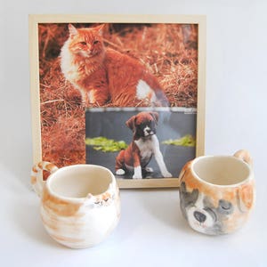 May include: Two ceramic mugs with animal faces. One mug features a brown and white cat face, and the other mug features a brown, white, and black dog face. The mugs are sitting on a white surface in front of a framed photo of a cat and a dog.