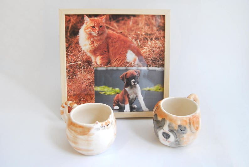 May include: Two ceramic mugs with animal faces. One mug features a brown and white cat face, and the other mug features a brown, white, and black dog face. The mugs are sitting on a white surface in front of a framed photo of a cat and a dog.