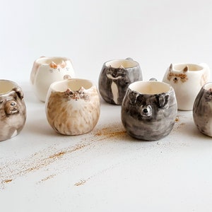 May include: Set of seven ceramic mugs featuring animal faces. The mugs are white with brown, black, and gray painted details. The animals include a cat, a dog, and a raccoon.