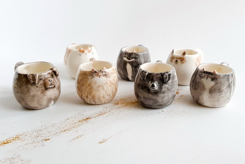 May include: Set of seven ceramic mugs featuring animal faces. The mugs are white with brown, black, and gray painted details. The animals include a cat, a dog, and a raccoon.