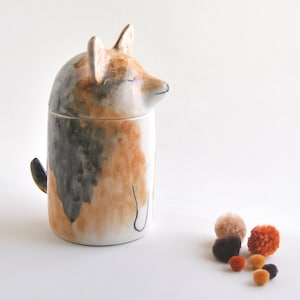 May include: A ceramic dog-shaped jar with a lid, painted in shades of brown, black, and white. The jar is sitting on a white surface with small, colorful pom-poms scattered in front of it.