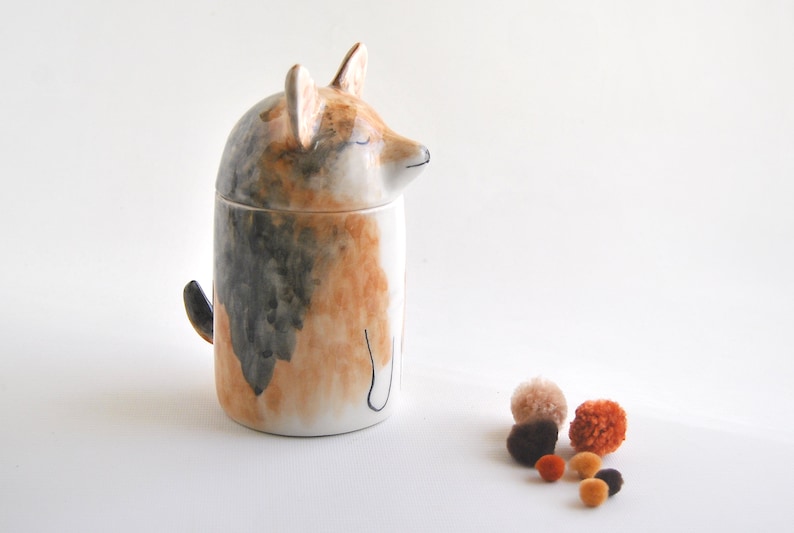 May include: A ceramic dog-shaped jar with a lid, painted in shades of brown, black, and white. The jar is sitting on a white surface with small, colorful pom-poms scattered in front of it.