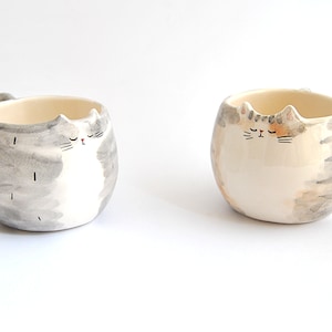 May include: Two ceramic mugs shaped like cats with gray and white fur and orange and white fur. The cats have closed eyes and are painted in a watercolor style.
