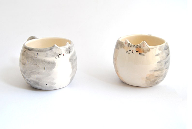 May include: Two ceramic mugs shaped like cats with gray and white fur and orange and white fur. The cats have closed eyes and are painted in a watercolor style.
