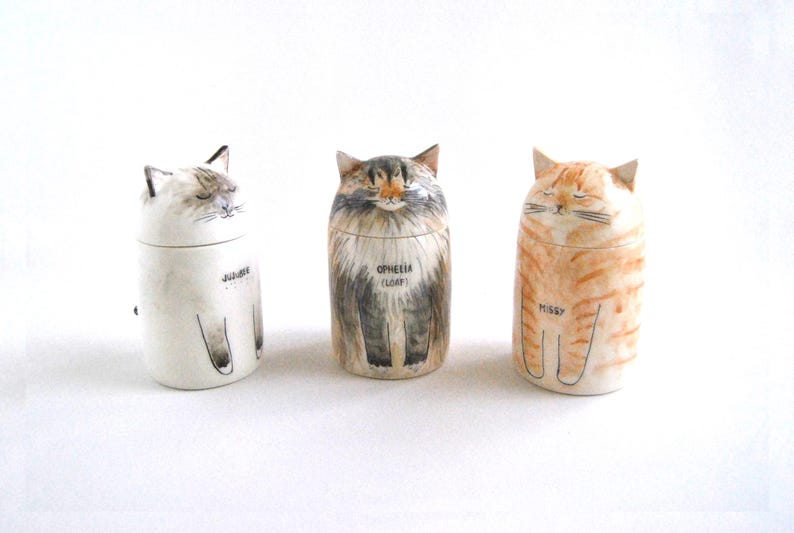 May include: Three ceramic cat-shaped containers with painted details. The containers are white, gray, and orange with black markings. The containers have the names "Julie", "Opheera (Loaf)", and "Missy" written on them.