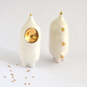 May include: Two white ceramic figurines with gold accents. One figurine has a large gold circle on its front and the other has three small gold circles. The figurines are standing on a white surface.