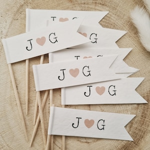 May include: A set of six white paper flags with wooden sticks. Each flag has a black letter J and G with a pink heart in between. The flags are arranged in a pile on a wooden surface.