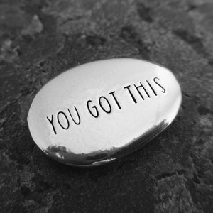 May include: A smooth, oval-shaped, silver-colored stone with the words "YOU GOT THIS" in black, sans-serif font. The stone is set against a dark, textured background, creating a high-contrast image.