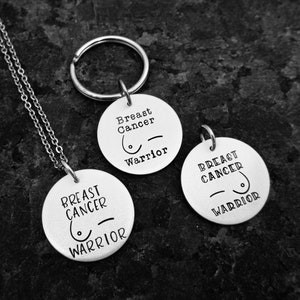 May include: Two silver keychains with a round charm that says "Breast Cancer Warrior" and a pink ribbon symbol. One keychain is attached to a silver chain necklace.