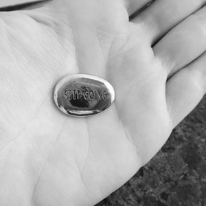 Pu&ograve; includere: Una piccola pietra ovale color argento con le parole incise "KEEP GOING". La pietra &egrave; tenuta in una mano aperta, con il testo ben visibile. L'immagine &egrave; in bianco e nero.