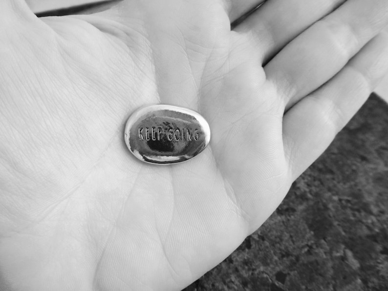 Pu&ograve; includere: Una piccola pietra ovale color argento con le parole incise "KEEP GOING". La pietra &egrave; tenuta in una mano aperta, con il testo ben visibile. L'immagine &egrave; in bianco e nero.