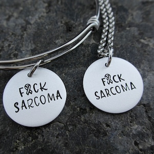 May include: A silver bracelet and necklace set with round silver charms that say "F*CK SARCOMA" with a ribbon symbol.