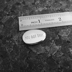 Pu&ograve; includere: Un piccolo ciondolo ovale in argento con la scritta "YOU GOT THIS". Un righello in acciaio inossidabile &egrave; posizionato accanto. Lo sfondo &egrave; una superficie strutturata grigio scuro. Il righello indica le dimensioni del ciondolo.