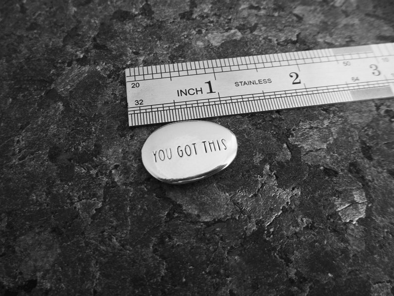Pu&ograve; includere: Un piccolo ciondolo ovale in argento con la scritta "YOU GOT THIS". Un righello in acciaio inossidabile &egrave; posizionato accanto. Lo sfondo &egrave; una superficie strutturata grigio scuro. Il righello indica le dimensioni del ciondolo.