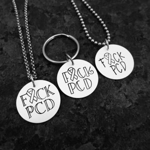 May include: Three silver necklaces with round pendants that say "F*CK PCD" with a ribbon symbol. The necklaces are on a dark background.