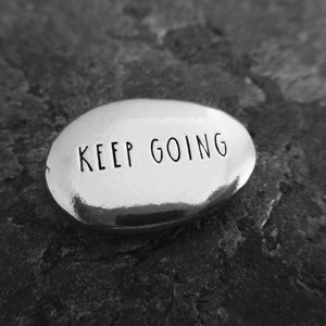 Pu&ograve; includere: Una pietra ovale argentata lucida con la scritta "KEEP GOING" incisa in un semplice font. La pietra poggia su una superficie strutturata grigio scuro, creando un contrasto tra la pietra liscia e lo sfondo ruvido.