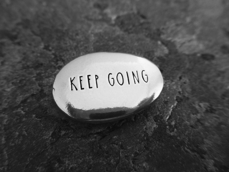 Pu&ograve; includere: Una pietra ovale argentata lucida con la scritta "KEEP GOING" incisa in un semplice font. La pietra poggia su una superficie strutturata grigio scuro, creando un contrasto tra la pietra liscia e lo sfondo ruvido.