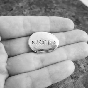 Pu&ograve; includere: Una pietra d'argento, di forma ovale, con la frase "YOU GOT THIS" incisa in maiuscolo. La pietra &egrave; tenuta in una mano aperta, con lo sfondo sfocato. L'immagine &egrave; in bianco e nero.