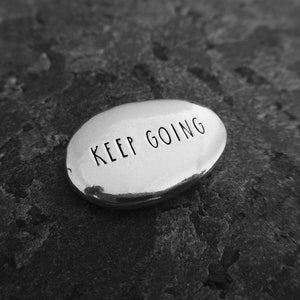 Pu&ograve; includere: Una pietra ovale color argento con le parole "KEEP GOING" incise in nero. La pietra &egrave; appoggiata su una superficie scura e strutturata. L'immagine &egrave; in bianco e nero.