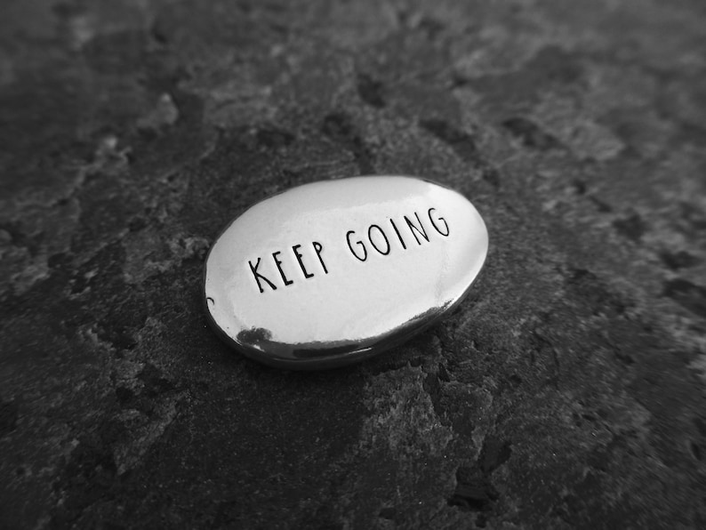 Pu&ograve; includere: Una pietra ovale color argento con le parole "KEEP GOING" incise in nero. La pietra &egrave; appoggiata su una superficie scura e strutturata. L'immagine &egrave; in bianco e nero.